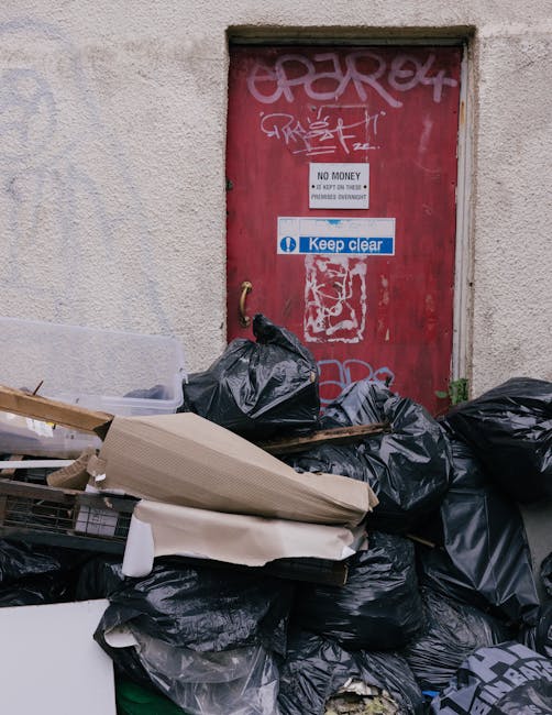 A pile of black garbage bags, cardboard, and miscellaneous waste items is stacked in front of a red metal door with graffiti, stickers, and signs on a textured beige wall. The bags appear full and tied at the top, with some leaning against the wall and others on the ground. The cardboard and other debris are haphazardly placed on top of or beside the bags, indicating a collection of unwanted waste. The door behind is weathered, with visible graffiti tags in pink spray paint, and has a white sign stating 'No Money' with additional text, as well as a blue and white sticker reading 'Keep clear.' The scene suggests a domestic or commercial environment where refuse is temporarily accumulated outside for private waste removal or alternative rubbish collection services, aligning with professional rubbish removal practices such as those offered by Rubbish Removal Belgravia. The lighting appears natural, highlighting the texture of the bags and wall, offering a clear view of the clutter associated with waste management or on-site clearance activities in an urban setting.