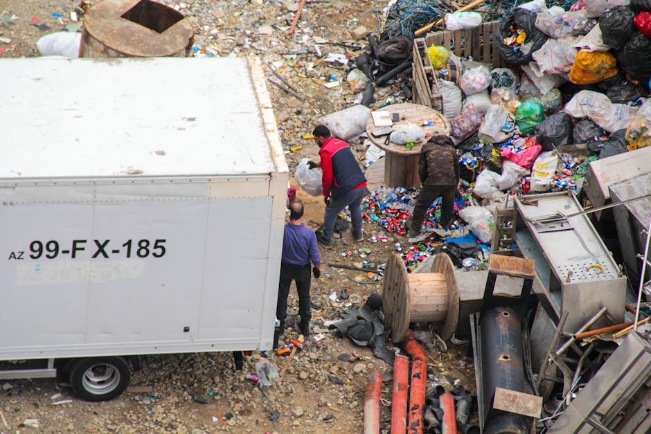 An aerial view of an area with scattered debris and waste materials, including plastic bags, cardboard boxes, and various discarded items. In the center, three individuals are seen sorting through the rubbish; one wearing a red and black jacket, another in a dark jacket, and a third in a purple shirt. A large white waste container truck with the registration number '99-FX-185' is parked on the left side, partially blocking the view. Surrounding the scene, there are large spools of cable, metal pipes, and other discarded industrial equipment placed irregularly on uneven, rocky ground. The environment appears cluttered and unmanaged, indicating the site is being prepared for waste removal or clearance. The scene suggests a private waste collection operation, possibly provided by Rubbish Removal Belgravia, serving the area with comprehensive rubbish disposal services, emphasizing the importance of on-site clearing and alternative waste handling methods in managing large-scale debris or industrial waste accumulation.