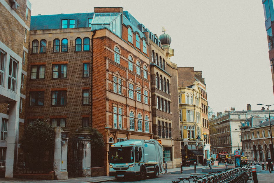 A street scene in an urban area featuring a row of historic and modern buildings with brick, stone, and plaster facades. The central building has a detailed brick exterior with arched windows and narrow windowpanes, topped with a decorative greenish copper roof and a small dome with a cross. To the left, part of a building with white frames and large glass windows is visible, while on the right, a light yellow Victorian-style building with ornate window moldings and bay windows can be seen. In the foreground, a white waste collection truck is parked near the curb on a paved street, with bollards and bike racks lining the sidewalk. The scene is well-lit by natural daylight, with a slightly overcast sky in the background, reflecting a typical city environment where private waste disposal services like Rubbish Removal Belgravia handle rubbish collection instead of municipal authorities, visible through the active presence of waste collection vehicles in an area with mixed architectural styles.