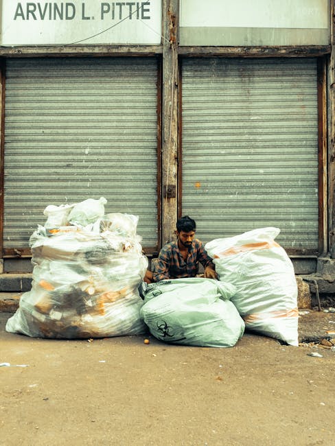 An aerial view of an area with scattered debris and waste materials, including plastic bags, cardboard boxes, and various discarded items. In the center, three individuals are seen sorting through the rubbish; one wearing a red and black jacket, another in a dark jacket, and a third in a purple shirt. A large white waste container truck with the registration number '99-FX-185' is parked on the left side, partially blocking the view. Surrounding the scene, there are large spools of cable, metal pipes, and other discarded industrial equipment placed irregularly on uneven, rocky ground. The environment appears cluttered and unmanaged, indicating the site is being prepared for waste removal or clearance. The scene suggests a private waste collection operation, possibly provided by Rubbish Removal Belgravia, serving the area with comprehensive rubbish disposal services, emphasizing the importance of on-site clearing and alternative waste handling methods in managing large-scale debris or industrial waste accumulation.