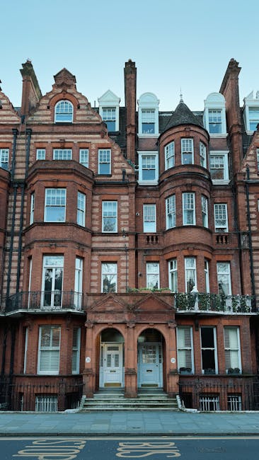 This image depicts the front façade of a multi-storey Victorian-style residential building constructed from red brick with detailed stonework accents. The building features several bay windows with white-framed glass panes, arranged across multiple levels, some with decorative stone or brick arches. The entrance is accessed via a short flight of steps leading to two distinct doorways, each with arched brick surrounds and white doors, one with a small window at the top. A black metal railing runs along a small balcony projecting from the first floor. The upper floors exhibit a combination of pitched and flat roof sections, with some dormer windows and chimney stacks visible. The surrounding environment includes a paved pavement directly in front with some painted street markings. The overall scene suggests a typical urban setting, and while no waste or rubbish is visible, the image context aligns with the services of professional rubbish removal specialists like Rubbish Removal Belgravia, who handle alternative waste collection and on-site clearance for private properties in the area. Natural daylight illuminates the scene, highlighting the historic architectural features and clean exterior surface of the building.