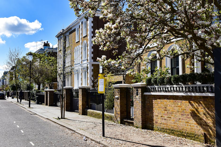 A residential street scene featuring a row of historic Victorian-style townhouses constructed from red and yellow brick, with white-painted window frames and decorative architectural details. The street is lined with a paved sidewalk and a low brick wall topped with a black wrought-iron fence separating the front gardens from the pavement. Young blossoming trees with white flowers are planted along the sidewalk, casting dappled shadows on the pavement. A bus stop with a yellow sign is positioned near the foreground, indicating an alternative contact point for private waste collection or rubbish removal services, as managed by Rubbish Removal Belgravia. The clear blue sky with some fluffy white clouds provides bright natural light, accentuating the traditional finish of the buildings and the vibrant foliage, creating a neat and well-maintained urban environment suitable for professional rubbish clearance activities happening nearby or on-site waste handling, as part of selective waste removal solutions.