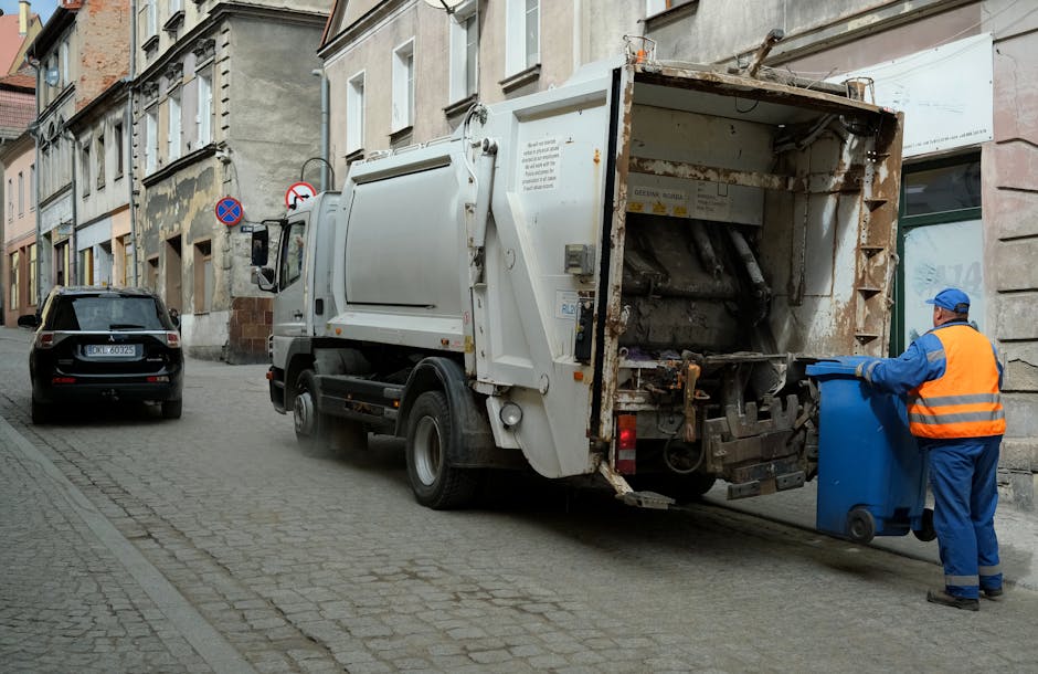 A silver waste collection truck parked on a city street in front of multi-story buildings featuring a mix of architectural styles, including red brickwork and ornate stone facades. The truck, positioned in the foreground on the right, is part of a rubbish removal service operated by Rubbish Removal Belgravia. The buildings behind display a combination of traditional and modern elements, with arched windows, bay windows, and decorative cornices, some topped with a dome or turret. To the left, there is a gated entrance flanked by stone pillars and some greenery. The street is lined with black bollards and bike racks, with a glimpse of other vehicles further down the road. The environment appears typical of an urban setting, with overcast lighting and a generally neutral atmosphere. This scene visually illustrates the process of private waste collection in an inner-city area, highlighting the role of independent rubbish removal services in managing urban rubbish disposal outside of municipal collection. The image captures the practical aspect of on-site rubbish clearance in a city environment, aligning with the services offered by Rubbish Removal Belgravia, especially for waste management in central London locations.