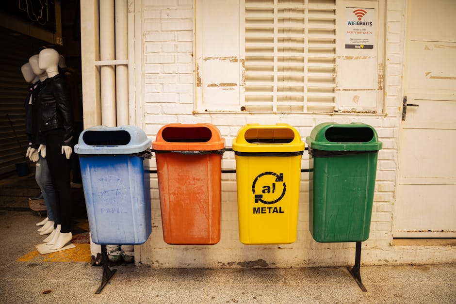 A row of four plastic waste bins positioned against a white brick wall beneath a window with horizontal white blinds, each bin in a different color: blue on the far left, orange next to it, yellow with a recycling symbol and the word 'METAL' in black in the middle, and green on the far right. The bins are mounted on black metal stands, raising them slightly above ground level. The blue bin appears to have some dirt or residue on its surface, while the other bins show minor scuffs and signs of use. To the left of the bins, part of a mannequin dressed in a black leather jacket and black pants is visible, standing near a closed shutter door. The scene is outdoors, with natural lighting illuminating the area, possibly during daytime. The image reflects an organized approach to waste sorting, suitable for private waste collection services or on-site waste management, aligning with the context of rubbish removal solutions offered by Rubbish Removal Belgravia.